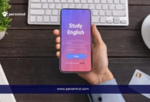 شخص يحمل هاتفًا ذكيًا تظهر عليه شاشة تطبيق بعنوان "Study English" كورس إنجليزي مجاني، فوق مكتب يحتوي على لوحة مفاتيح ودفتر وفنجان قهوة.