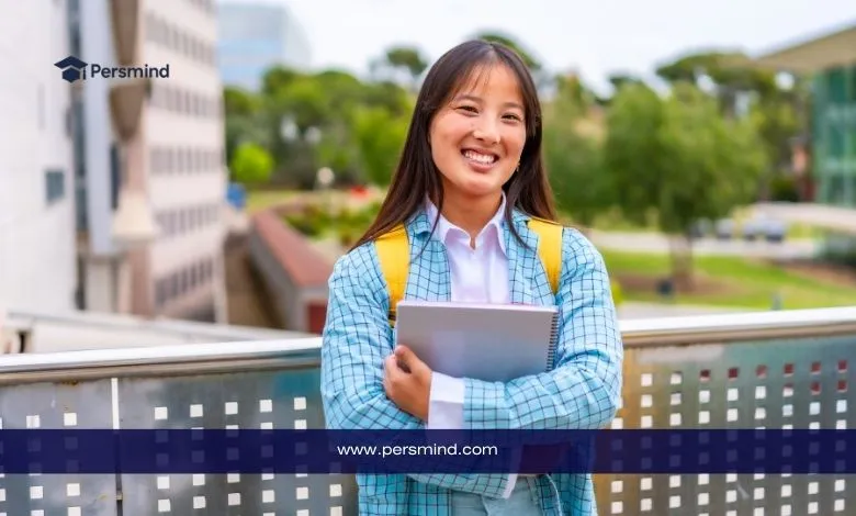 طالبة تحمل كتبًا وتقف في حرم جامعي خارجي مبتسمـة أمام الكاميرا.