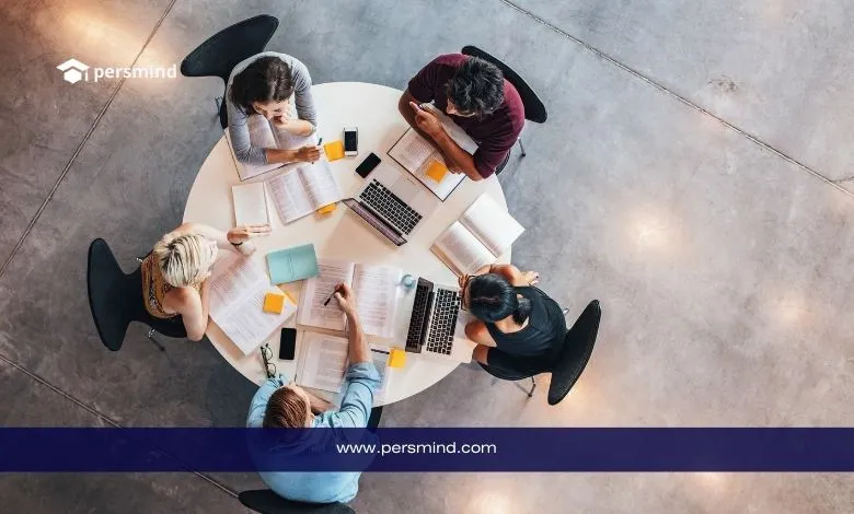 طلاب يجلسون حول طاولة مستديرة يعملون معًا على دراسة جماعية باستخدام الكتب وأجهزة الحاسوب المحمولة.