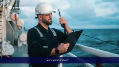 مهندس بحري يرتدي خوذة سلامة على متن سفينة أثناء فحص المعدات البحرية باستخدام جهاز اتصال ولوح بيانات، في سياق دراسة الهندسة البحرية.